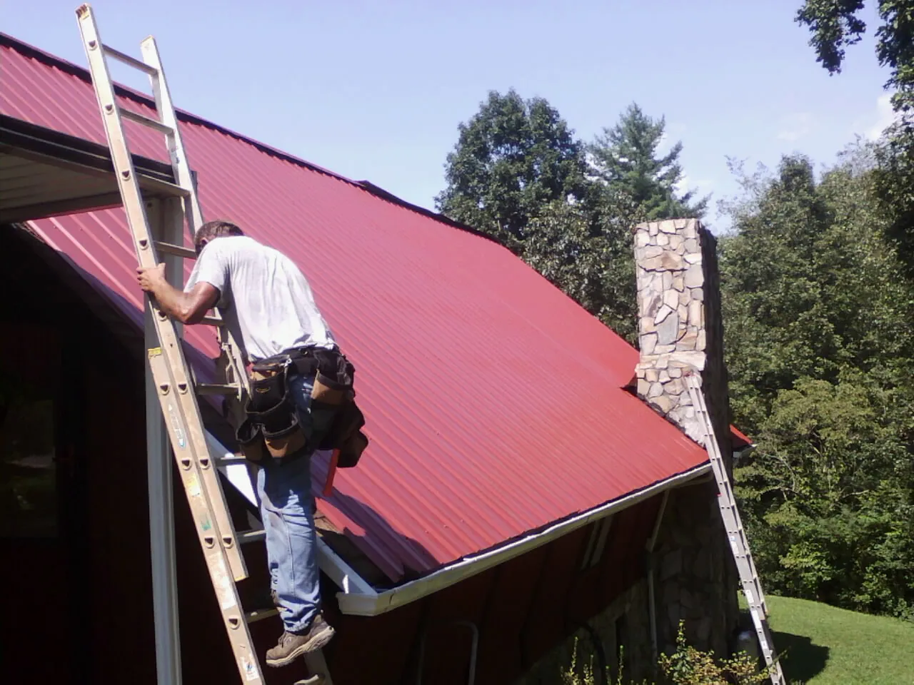 Roofer on a ladder beside a red metal roof and stone chimney — Soffit & Fascia Repair by Roofing Brothers in Sisters